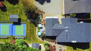 An aerial view of a residential area with a tennis court, a covered patio area, and several houses. - Bayfront PNW Lodge: Dock, Pickleball & Guest House (Bainbridge Island)