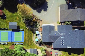 Vue aérienne d’un quartier résidentiel avec un court de tennis, un patio couvert et plusieurs maisons.