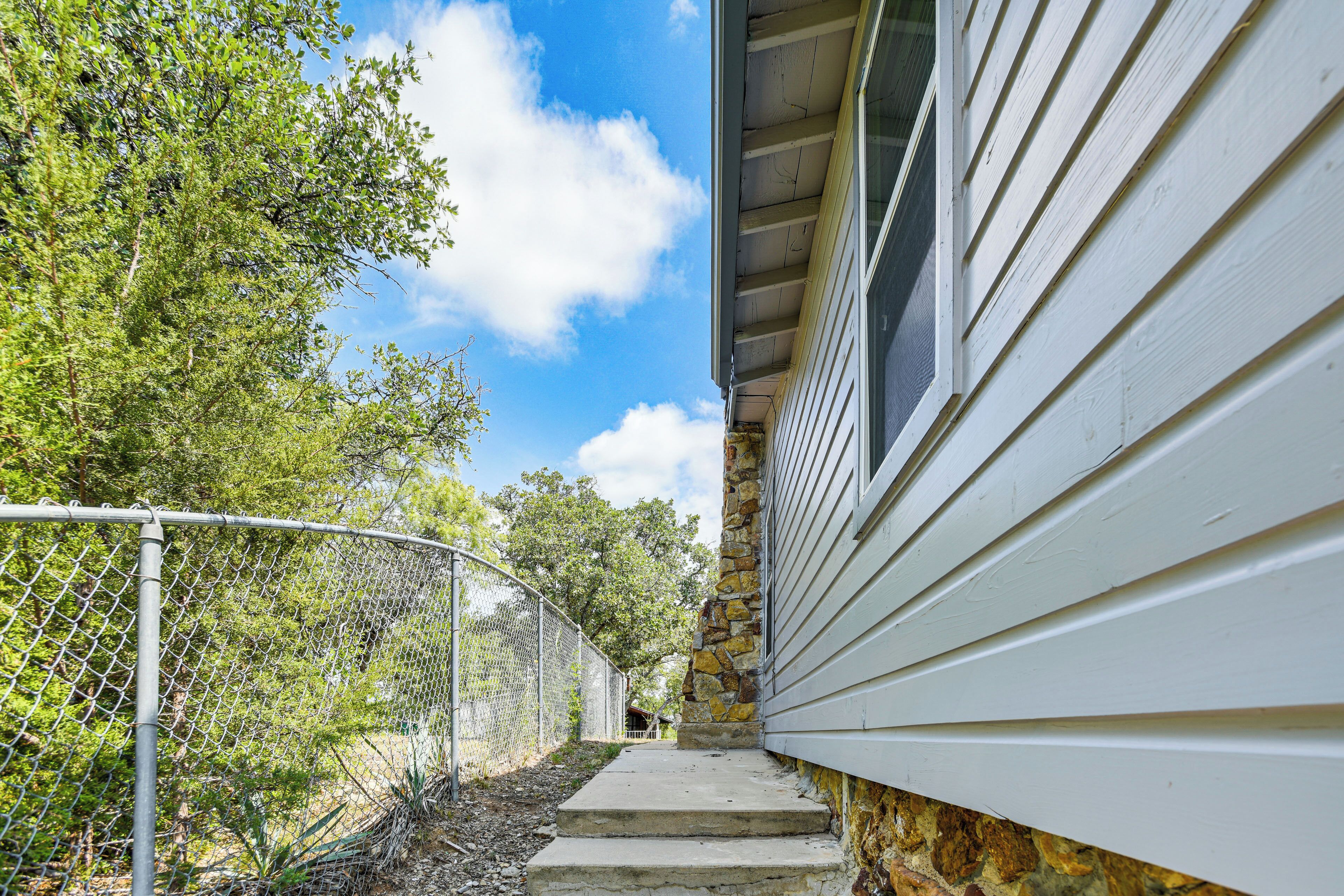 A pathway leading to a house with a stone pillar, a chain-link fence, and trees in the background.