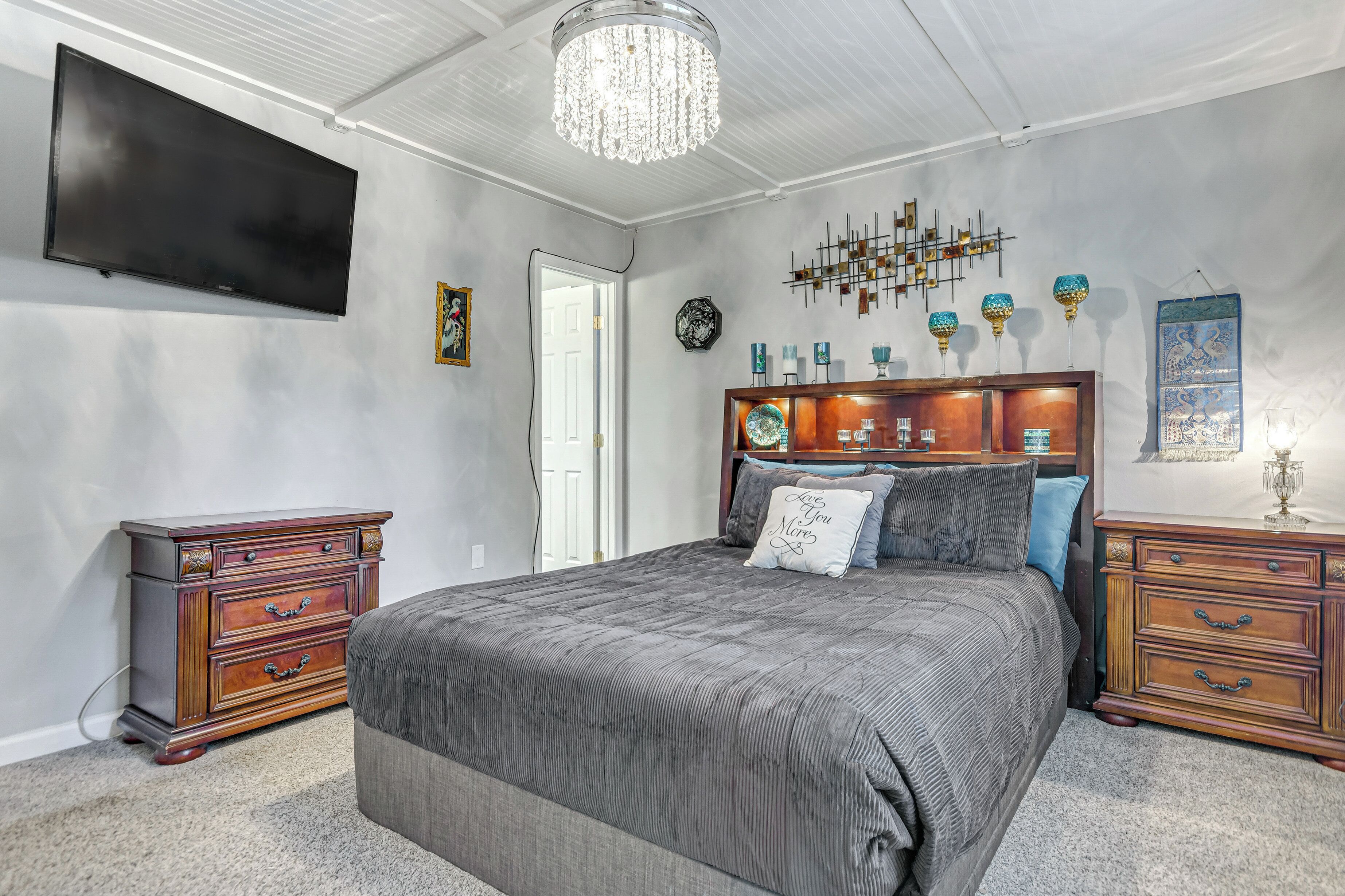 A bedroom with a large bed, a TV mounted on the wall, a chandelier, and a wooden dresser.