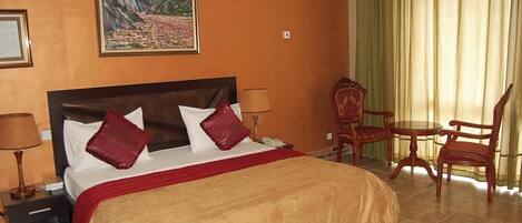 A hotel room with a large bed, a painting of a mountain landscape, a chair, and a table.
