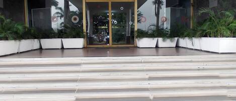 A building entrance with a glass door, a staircase leading up, and potted plants on either side.