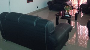 Un salon avec un canapé noir, deux fauteuils, une table basse ornée d’un vase et un téléviseur à écran plat fixé au mur.