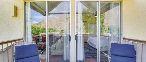 A covered patio with two blue lounge chairs, a sliding glass door leading to a patio area with a bed and a table, and a view of a mountainous landscape.