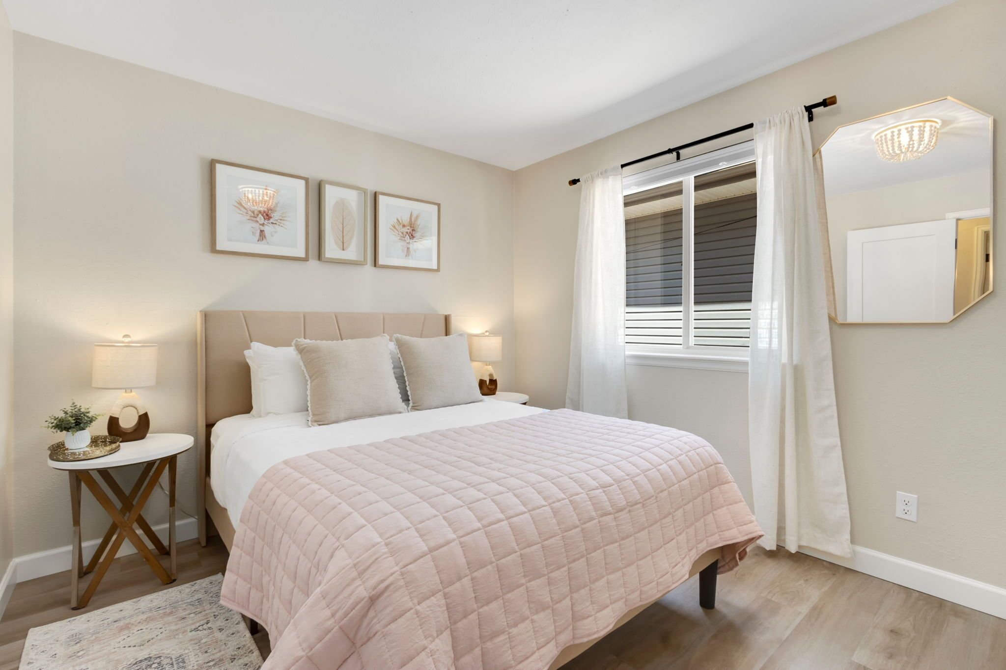 A bedroom with a bed, bedside tables, a nightstand, a window with curtains, and wall-mounted artwork.