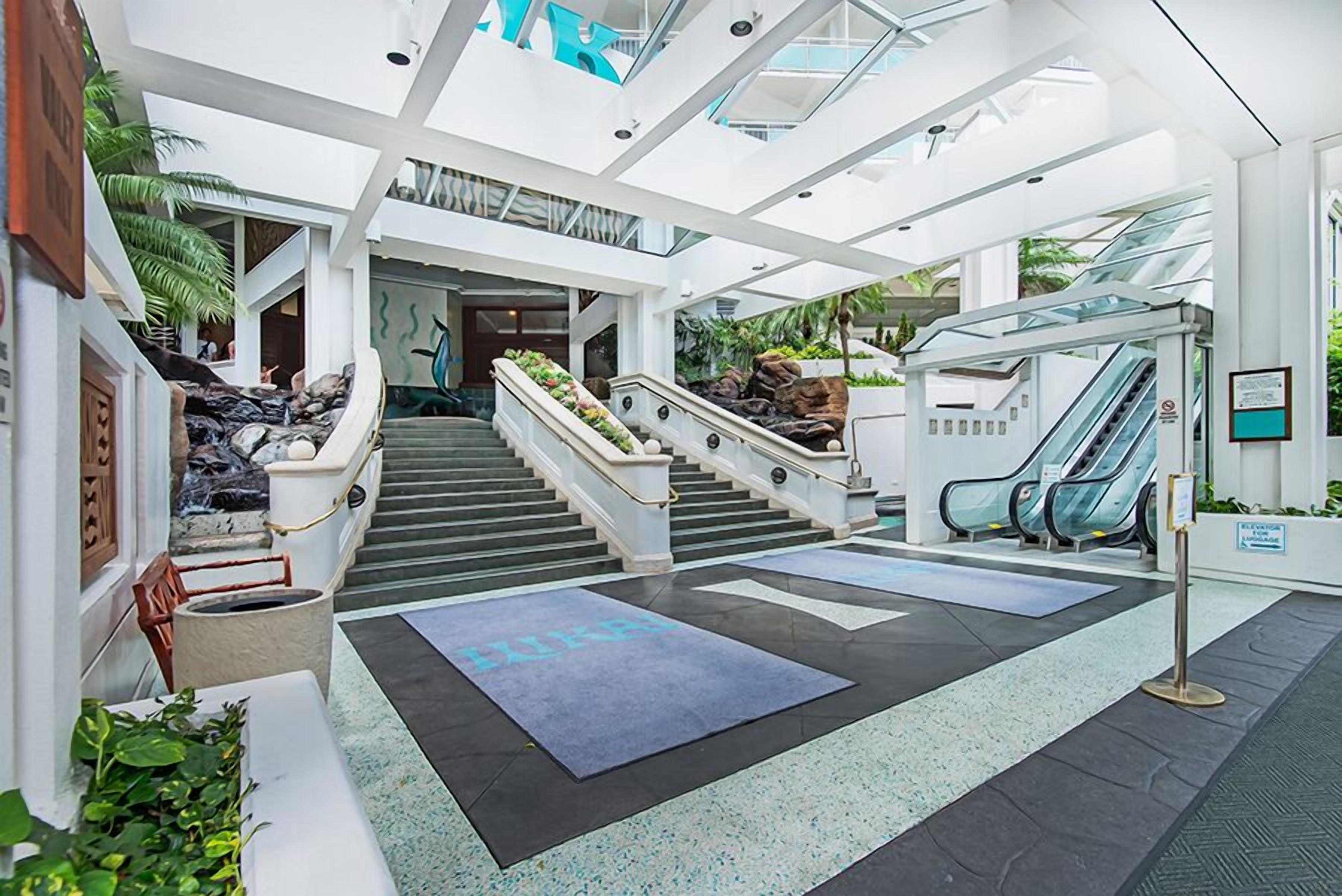 A modern indoor atrium with a central staircase, escalators, and a seating area with a blue mat.