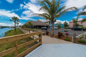 A wooden deck leading to a building with a thatched roof, a palm tree, and a view of the water.
