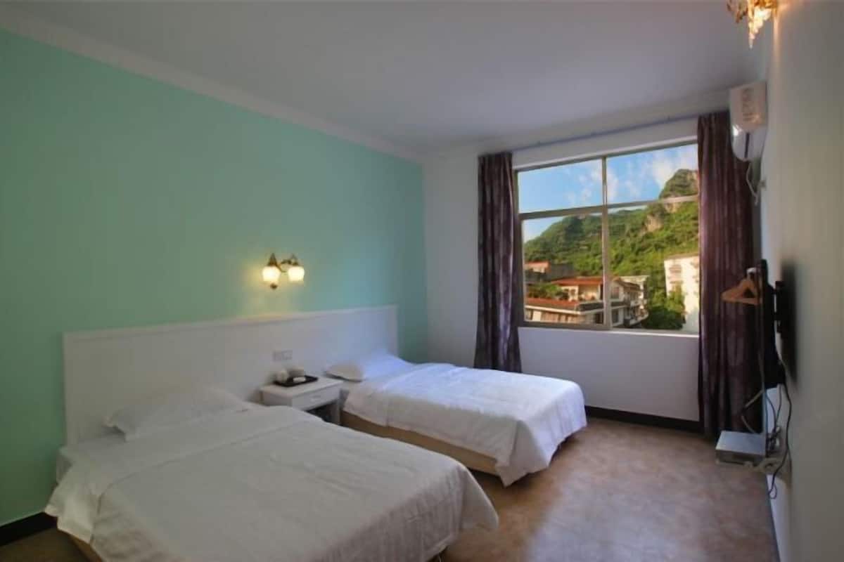 A room with two beds, a window with a view of buildings, and a wall-mounted light fixture.