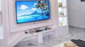 A living room with a flat-screen TV mounted on a white wall, a black coffee table, and a marble floor. - MONDEESTARS LUXURY HOME (Ibadan)