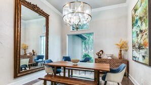 Un comedor con una gran mesa de madera, sillas tapizadas en azul, un banco, una araña, un espejo grande y un cuadro en la pared.