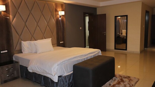 A modern bedroom with a large bed, a nightstand, a bench, and a mirror. - Delad Hotel and Suites (Ibadan)