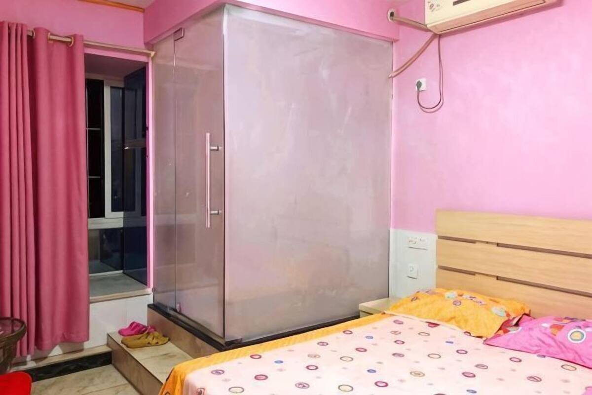 A bedroom with a wardrobe, a bed with a patterned bedspread, a window with pink curtains, and a wall-mounted air conditioner.
