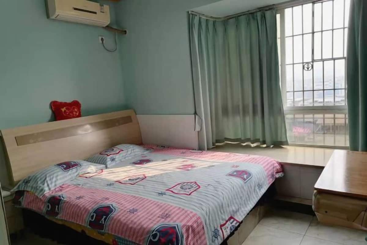 A bedroom with a bed, a window with curtains, a bedside table, and a wall-mounted air conditioner.