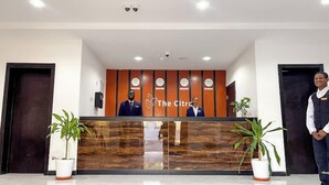 A reception desk with a logo, two clocks, and a security camera. There are potted plants and a man standing to the right. - The Citron Hotels (Ibadan)