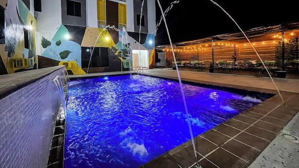 A swimming pool with a water fountain, surrounded by a building with yellow and white exterior walls and a covered outdoor seating area with string lights. - The Citron Hotels (Ibadan)