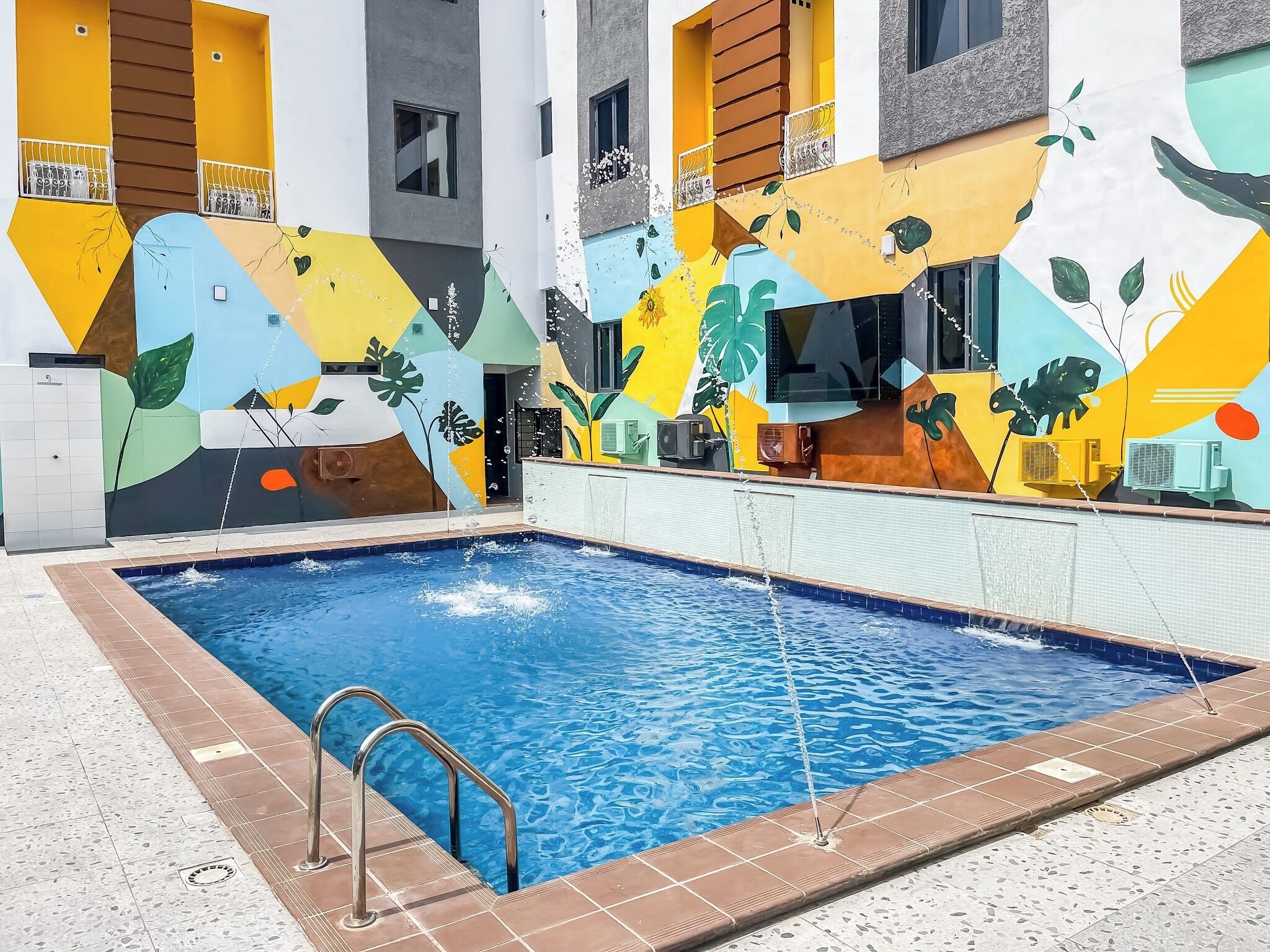 A swimming pool with a tiled deck and metal railings, surrounded by a building with colorful murals of plants and geometric shapes.