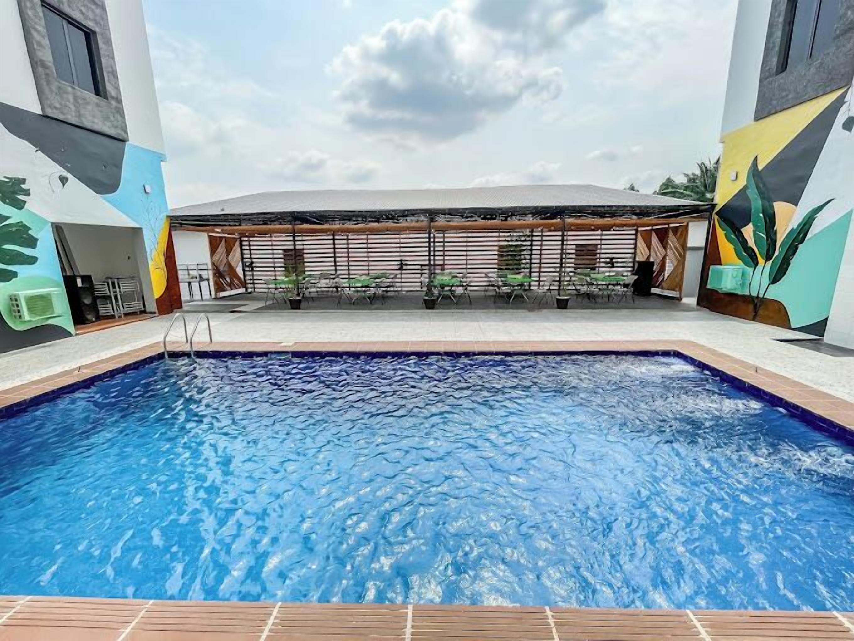 A swimming pool with a wooden deck, surrounded by a covered seating area with chairs and tables, and a building with colorful murals.