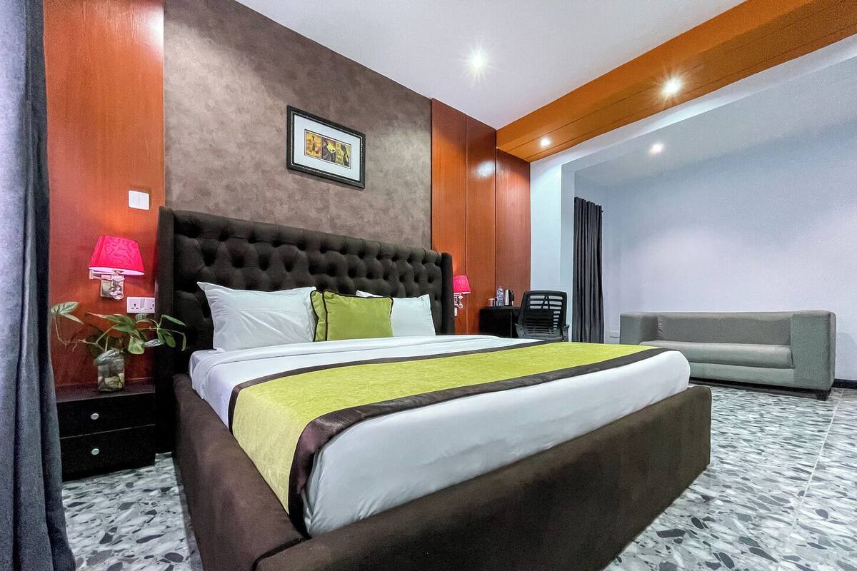 A hotel room with a large bed, a grey sofa, a red lamp, and a framed picture on the wall.