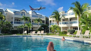 Une vue sur la piscine où l’on aperçoit une personne se détendant, un ciel bleu dégagé et un avion qui vole au-dessus.