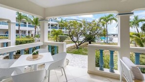 Un espace extérieur abrité, avec une table et des chaises blanches, une petite plante, et une vue sur une piscine et des bâtiments en arrière-plan.