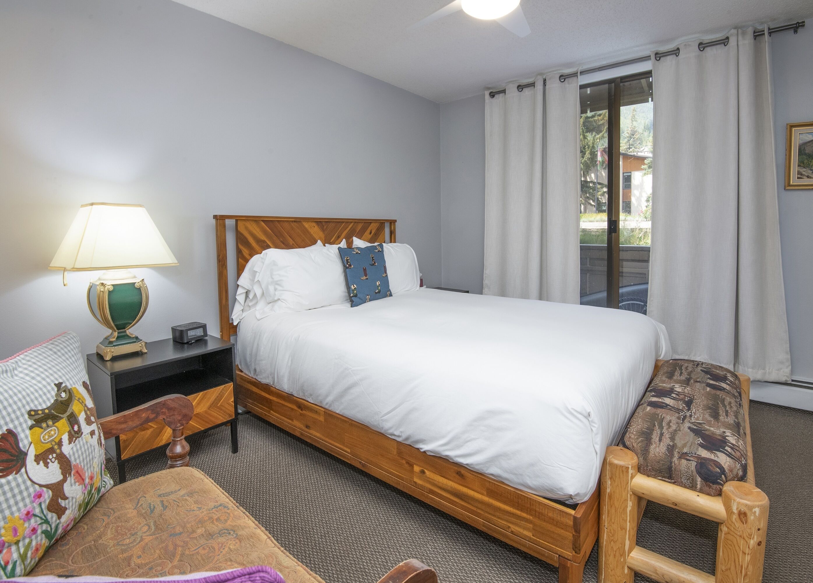 A neatly arranged hotel room with a wooden bed, a bedside table with a lamp, a chair, and a view of the outdoors through a window.