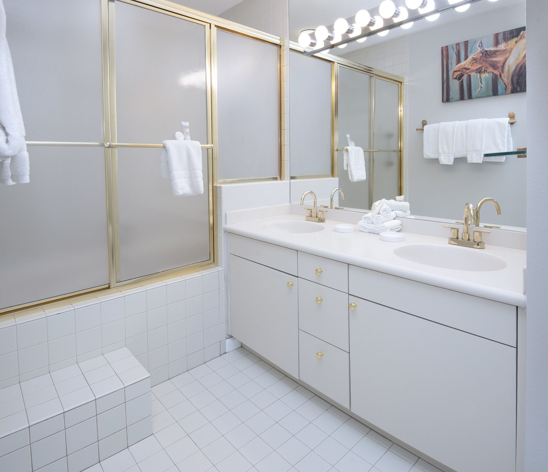 A modern bathroom with two sinks, gold fixtures, and a large mirror with lights above.