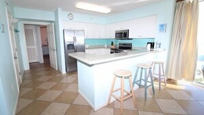 Cocina moderna con una isla central, armarios blancos y un microondas. Hay dos taburetes en la isla y un refrigerador a la izquierda.