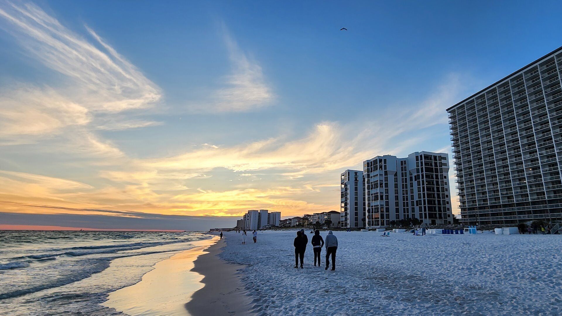 A beach with a row of high-rise buildings, a clear sky, and a few people walking.