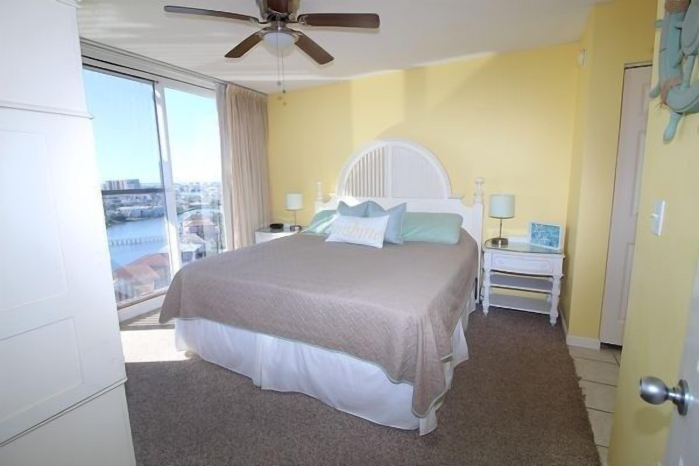 A hotel room with a bed, a ceiling fan, a nightstand, and a view of a cityscape through a large window.