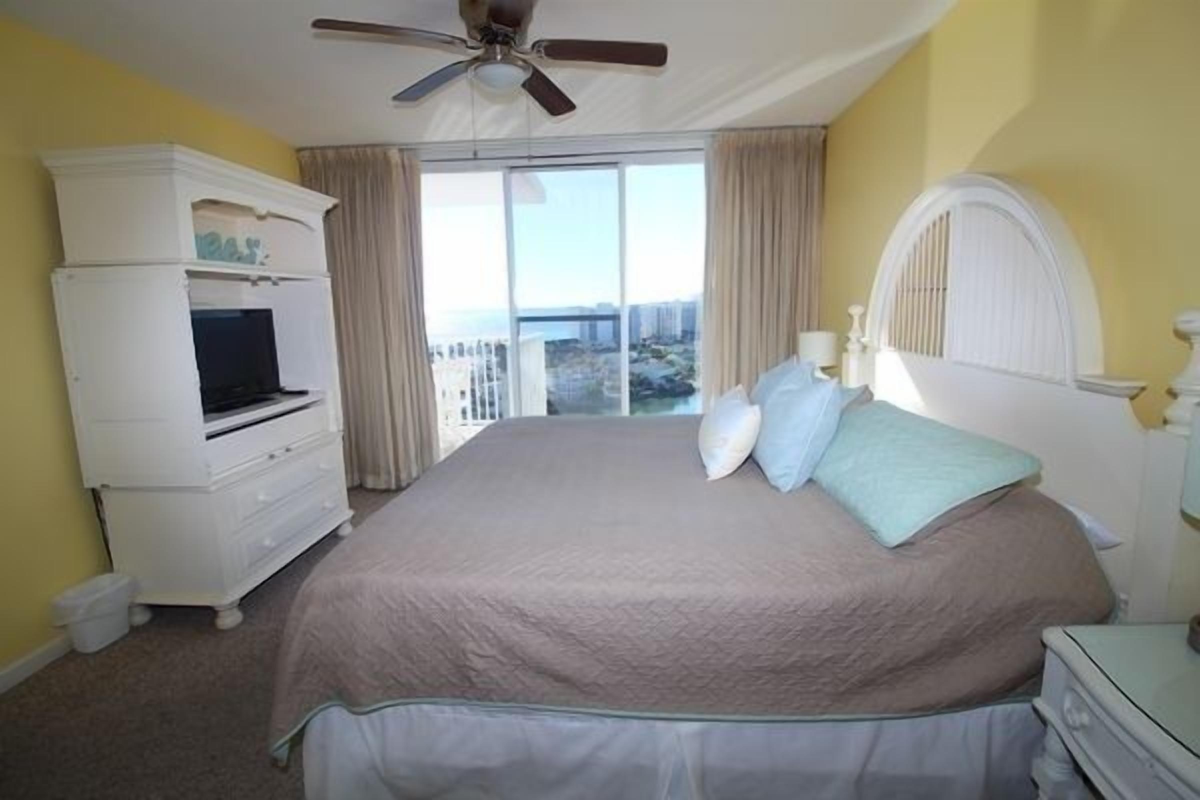A hotel room with a bed, a TV, a ceiling fan, and a view of the city.
