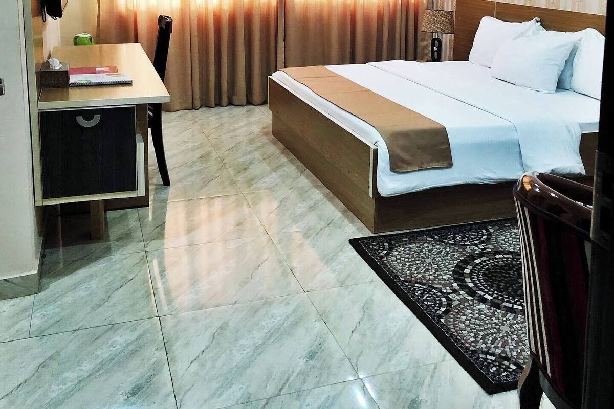 A hotel room with a bed, a desk, a chair, a small table, a decorative vase, and a patterned rug.