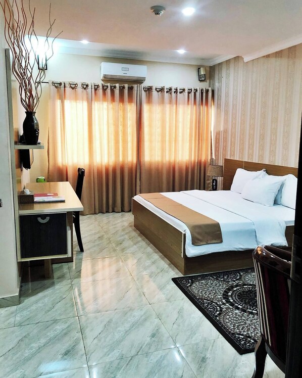 Habitación de hotel con cama, escritorio, silla, mesita pequeña, un jarrón decorativo y alfombra estampada.