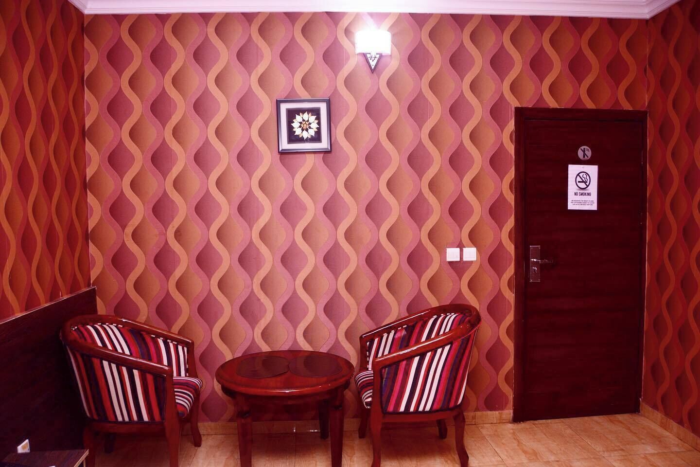 Une pièce avec un papier peint à motifs, un tableau encadré accroché au mur, une porte sur laquelle est apposée une pancarte « Interdiction de fumer », et deux chaises à rayures disposées autour d’une petite table.