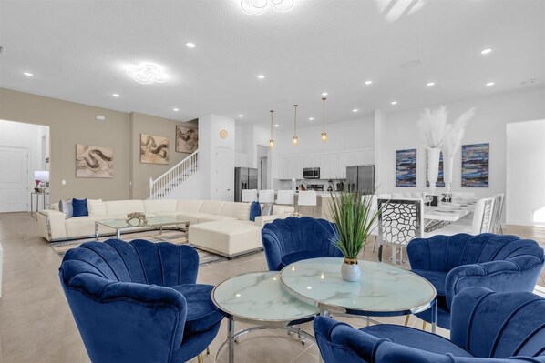 Un vaste espace de vie avec une cuisine moderne en arrière-plan, un coin repas avec une table et des chaises blanches, et un escalier menant à un autre niveau.