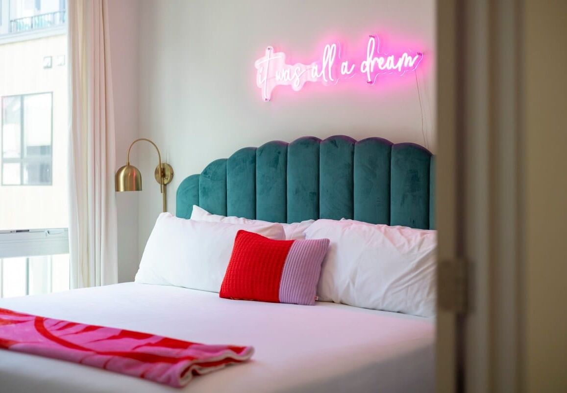 Una cama bien hecha con un cabecero color turquesa, almohadas blancas y rojas, y un letrero de neón que dice "Twice a dream".