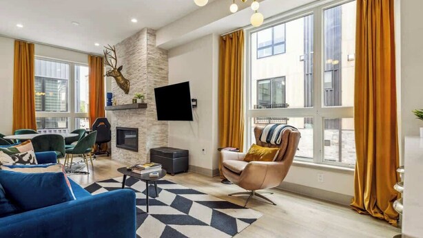 A modern living room with a stone fireplace, a mounted TV, and a large window with curtains. - Roof Deck | MTN View | Hot Tub & Cold Plunge (Silverthorne)