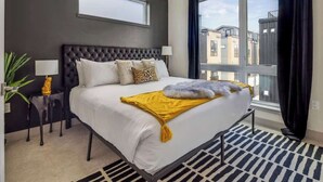 A modern hotel room with a large bed, a view of buildings through the window, and a small side table with a lamp. - Roof Deck | MTN View | Hot Tub & Cold Plunge (Silverthorne)