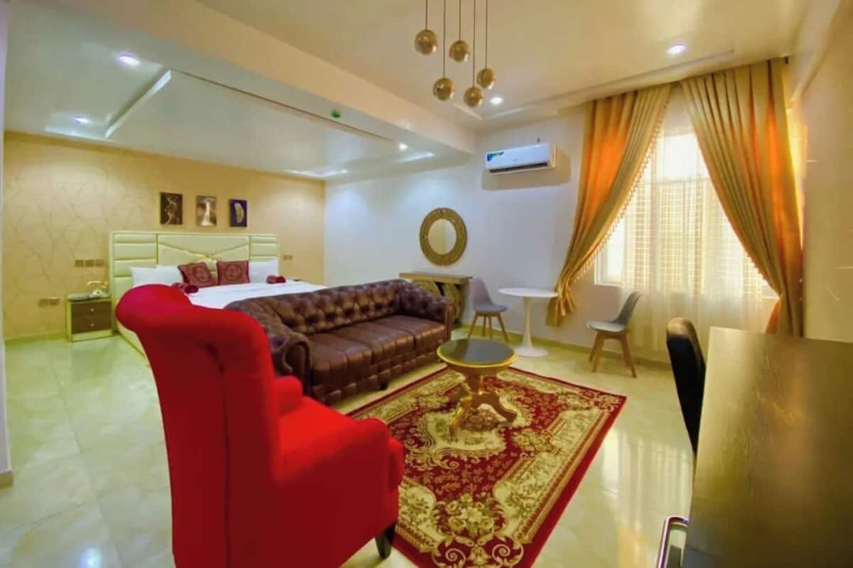 A hotel room with a bed, a red armchair, a brown sofa, a round coffee table, and a dining table with chairs.
