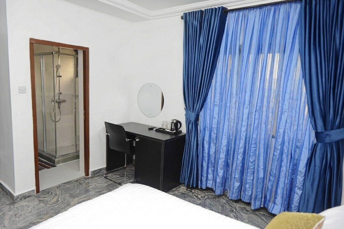 A hotel room with a shower, a desk, a chair, a bed, and blue curtains.