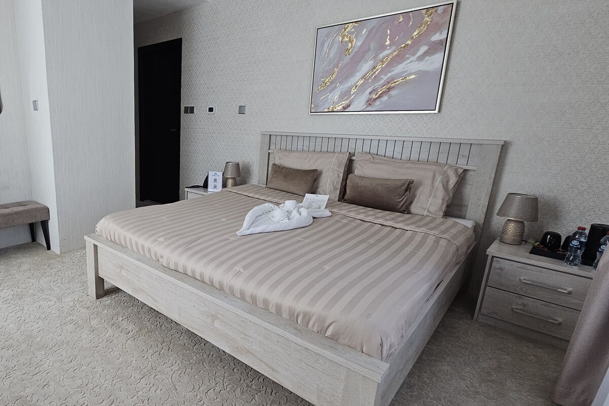 A hotel room with a large bed, a desk, a chair, a bench, a bedside table, and a wall-mounted mirror.