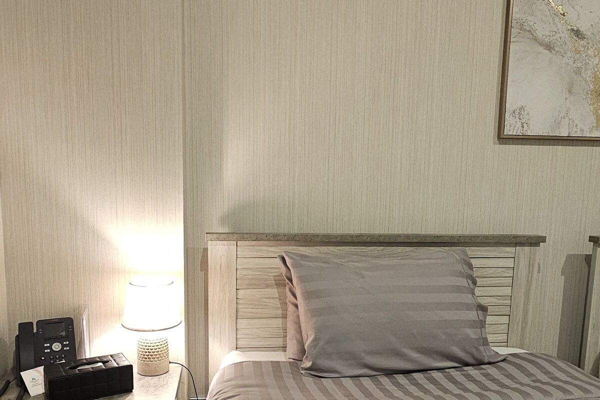 Camera d'albergo con un letto, un comodino con una lampada, un telefono e un asciugamano decorativo sul letto.