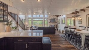 A spacious kitchen with dark cabinetry, a central island, and a dining area with a table and chairs. There is a staircase leading to an upper level, and large windows provide natural light and views of the outdoors.