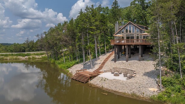 A lakeside cabin with a wooden deck, a stone fire pit, and a view of the lake.