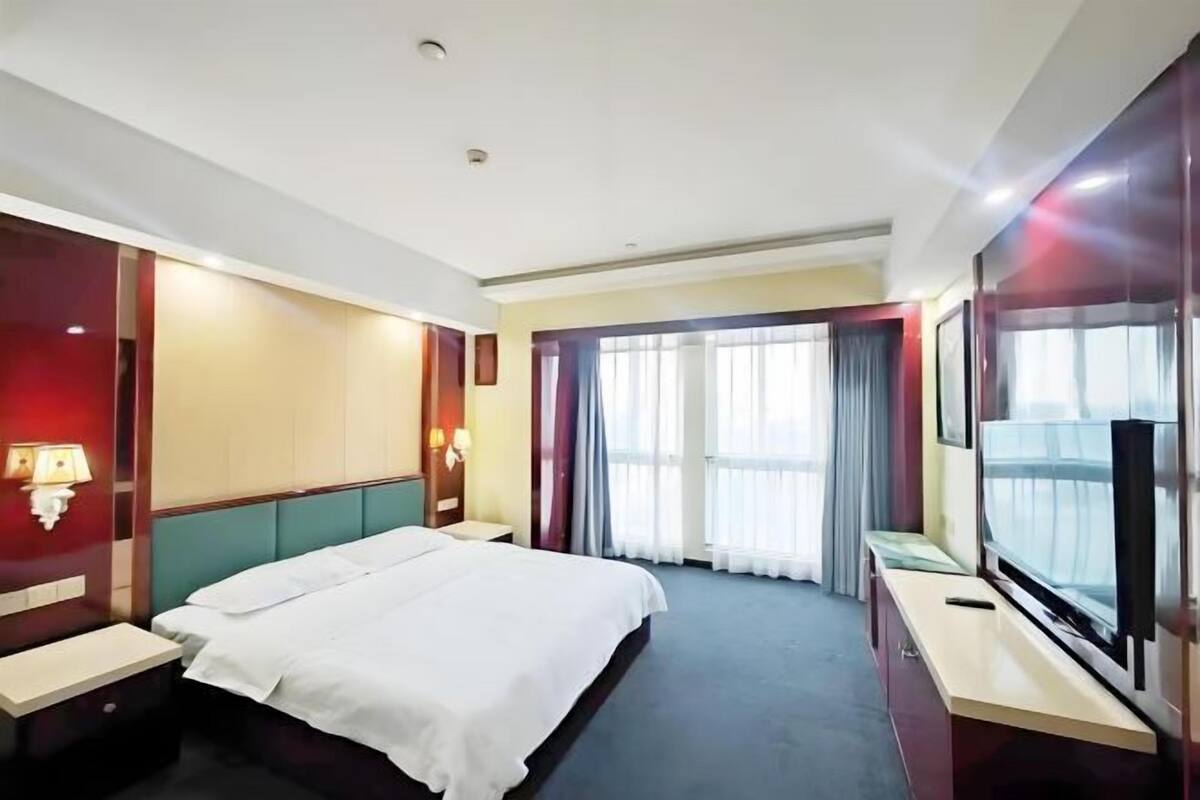 A hotel room with a large bed, a TV, a desk, and a window with curtains.
