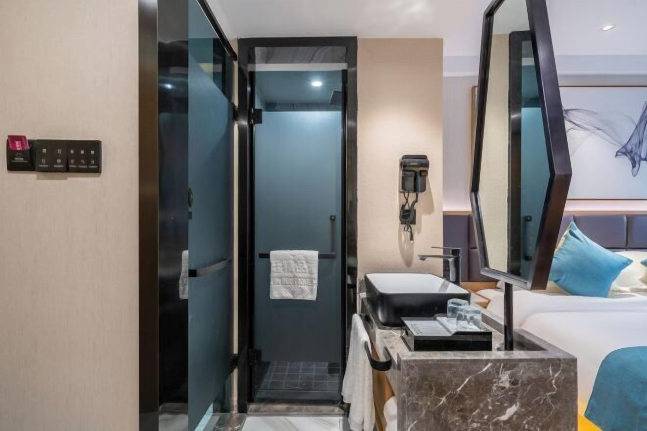A hotel room with a glass-enclosed shower, a sink, a mirror, a bed with blue pillows, and a wall-mounted phone.