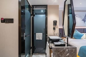 Quarto de hotel com chuveiro em gabinete de vidro, pia, espelho, cama com almofadas azuis e telefone fixado na parede.