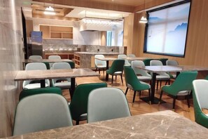 Een moderne eetruimte met marmeren tafels en groene stoelen, een keuken op de achtergrond en een projectiescherm met een landschap.