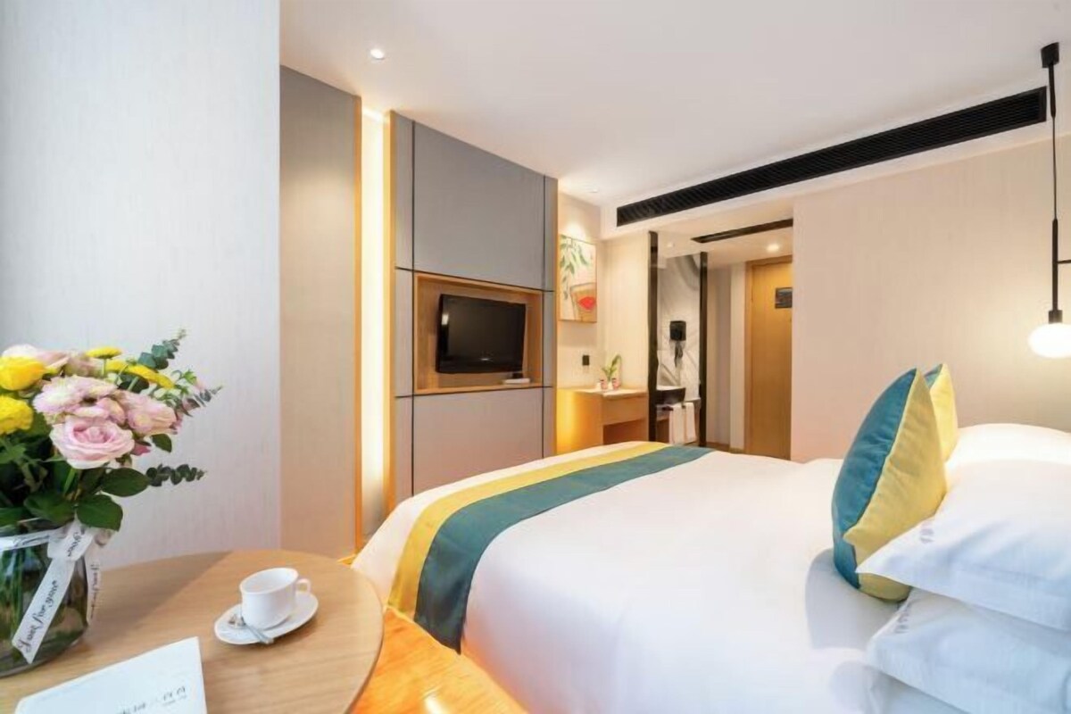 A hotel room with a bed, a TV, a desk with a cup and saucer, and a vase of flowers.