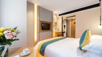 A hotel room with a bed, a TV, a desk with a cup and saucer, and a vase of flowers.