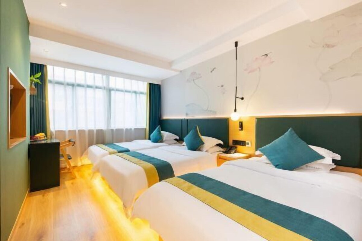 A hotel room with two beds, a large window, a wall with floral artwork, and a bedside table with a lamp.